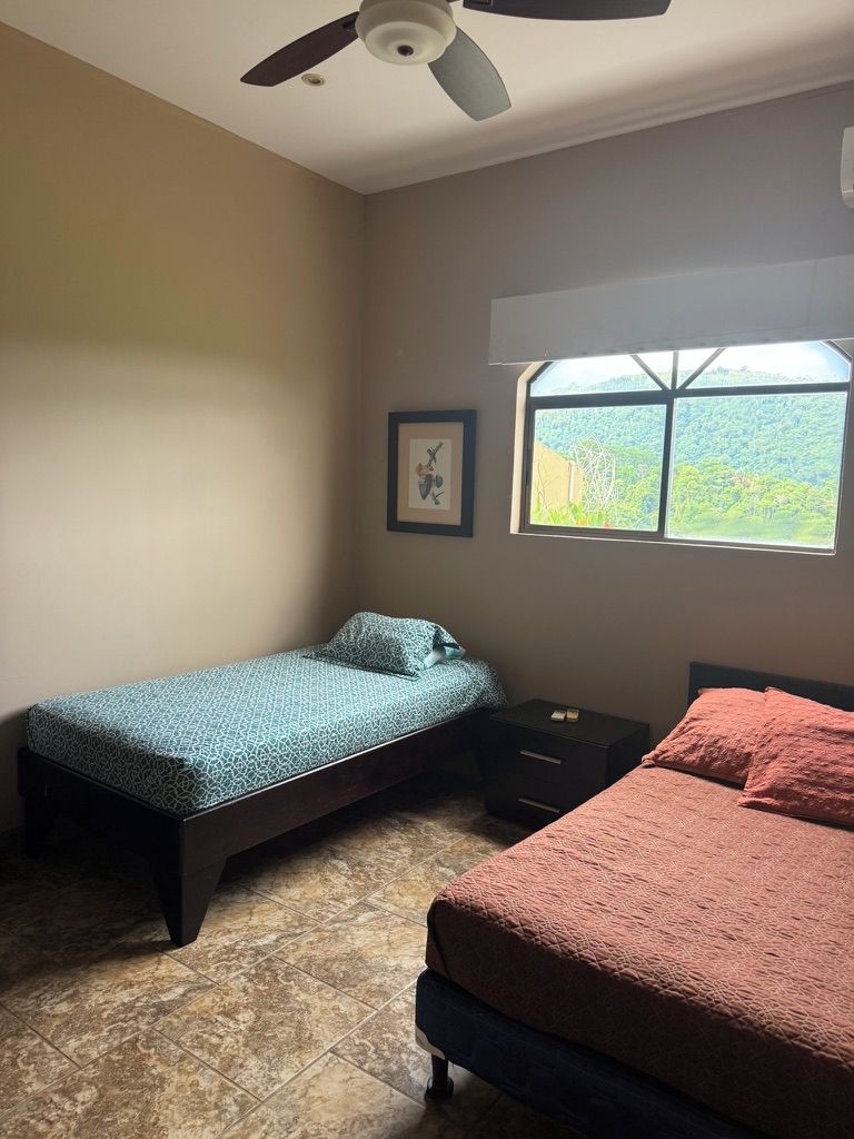 A bedroom with two beds, a ceiling fan, a window with a view, and a framed picture on the wall.