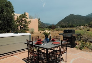 Un área de comedor al aire libre con una mesa dispuesta para cuatro personas, rodeada de sillas, una parrilla y un jacuzzi.
