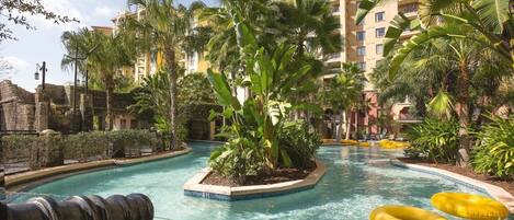 Zona de piscina con palmeras, un muro de piedra y un edificio al fondo.