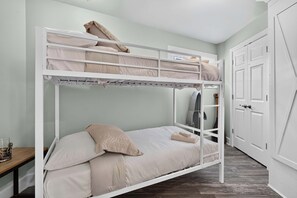Una cama litera con una cama inferior y una cama individual, ambas con ropa de cama color beige. Hay un banco de madera y una puerta con manija a la derecha.