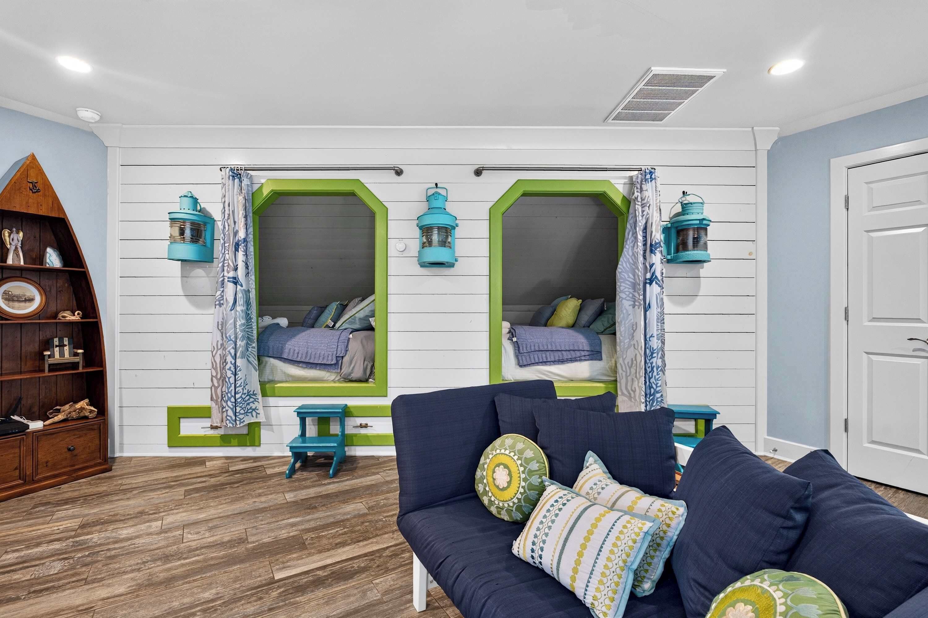 Een kamer met een houten vloer, een blauwe bank met gekleurde kussens, en twee ramen met groene lijst en gordijnen.