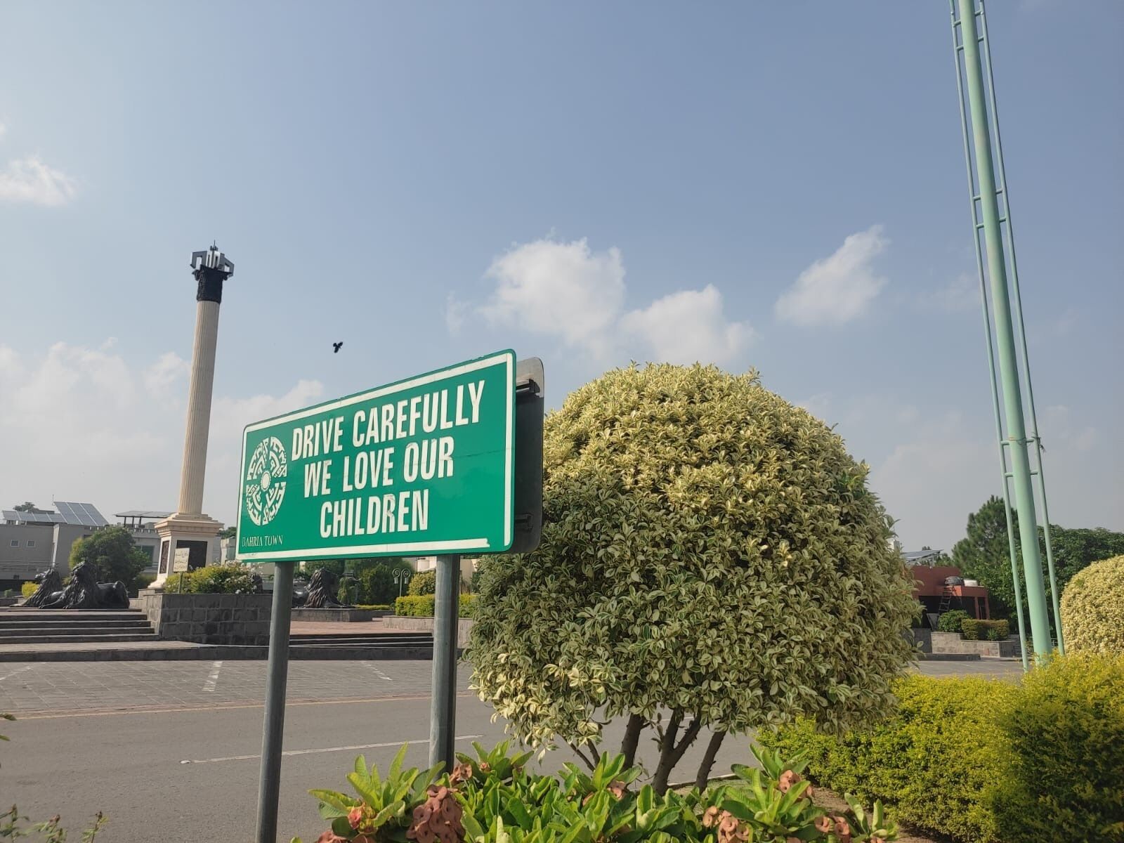 A green sign with white text in a public space, surrounded by greenery and a monument.