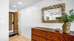 Una habitación con un aparador de madera, un espejo con marco de concha marina, una planta en maceta y una puerta que da a otra habitación.