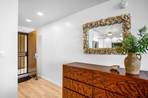 A room with a wooden dresser, a mirror with a shell frame, a potted plant, and a door leading to another room. - Valley Isle Villas | Oceanfront Studio Sleeps 4 | Car Incl. w/ 6+ Nights | VIR-107 by KBM (Lahaina)
