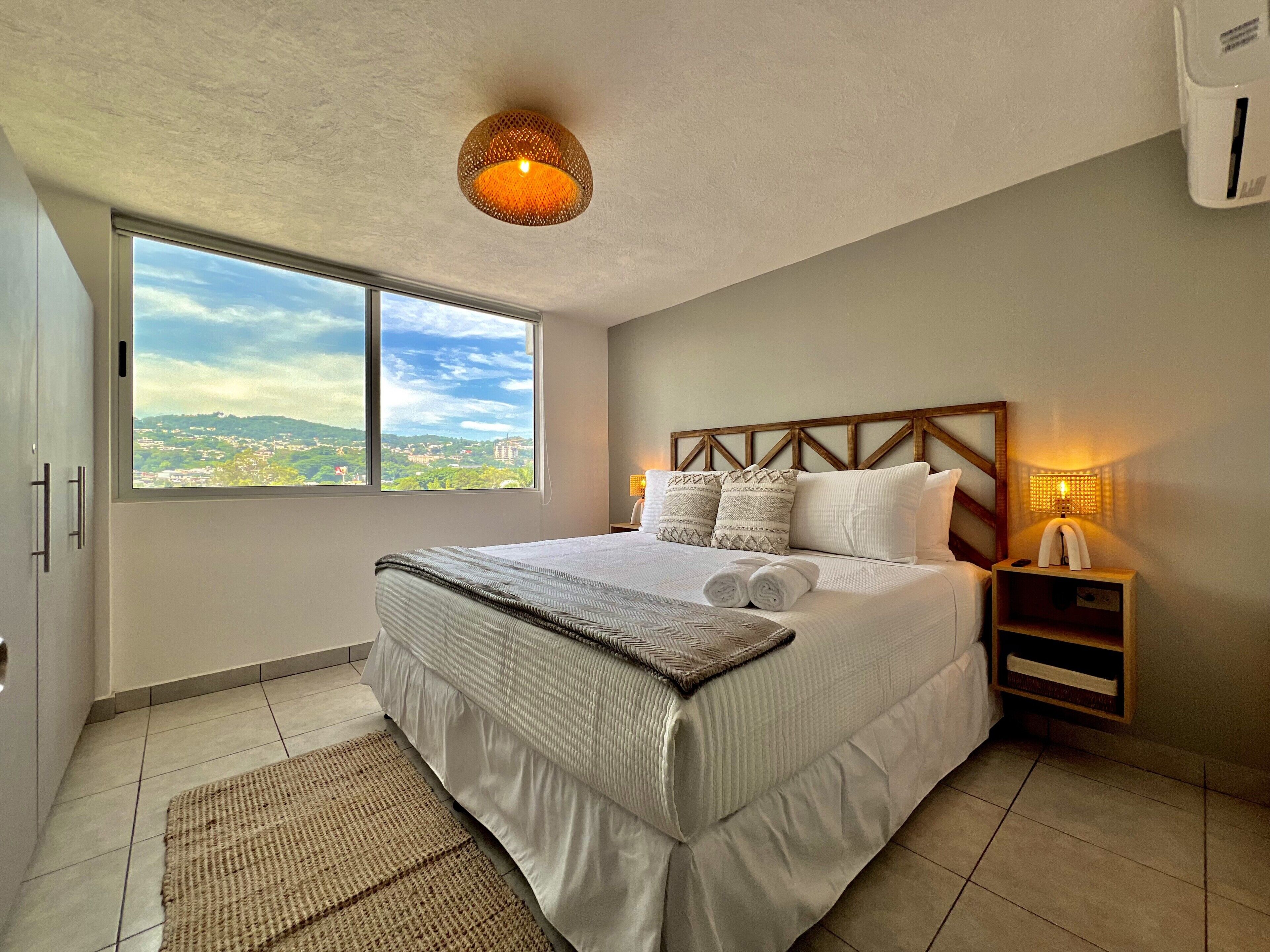 A bedroom with a large bed, a view of a cityscape through a window, a ceiling light fixture, and a bedside table with a lamp.