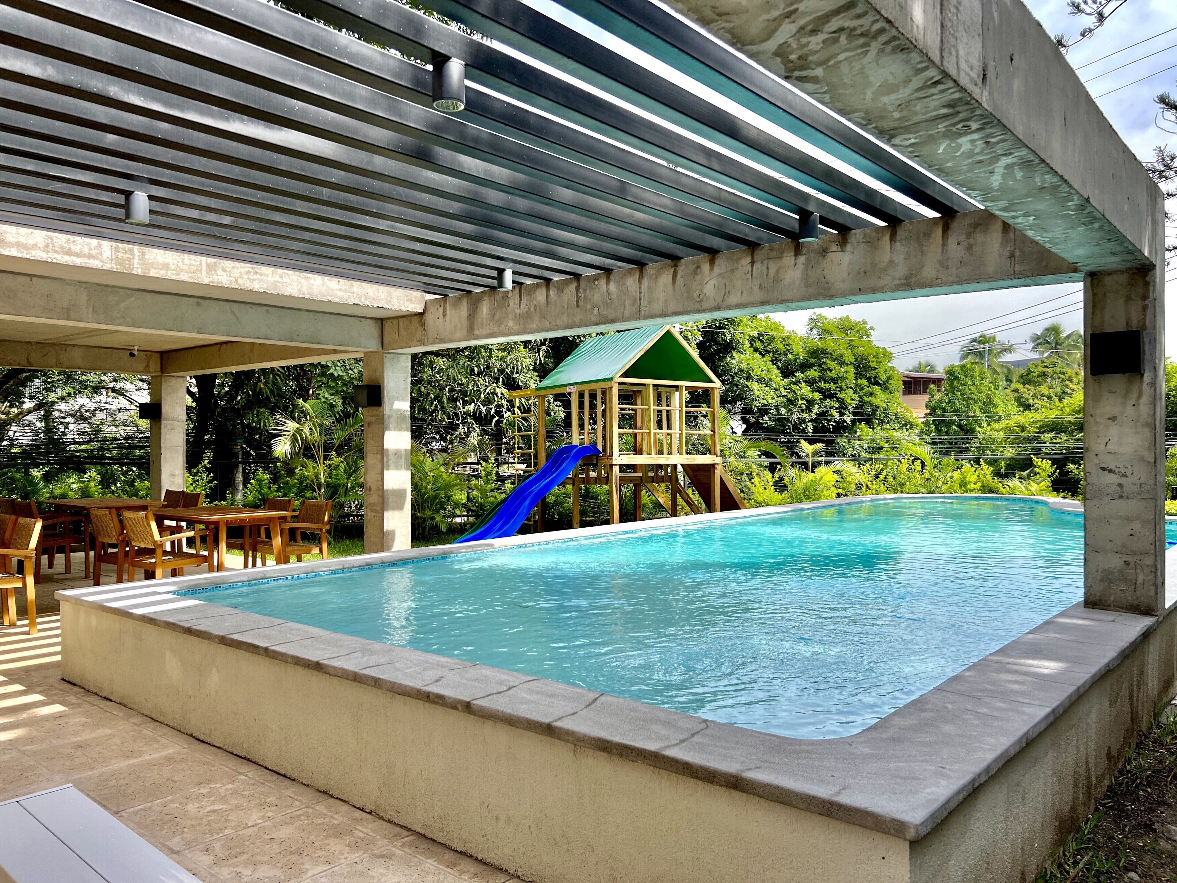 An outdoor swimming pool with a slide, surrounded by chairs and a covered area.