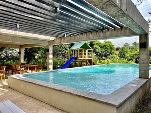 An outdoor swimming pool with a slide, surrounded by chairs and a covered area.