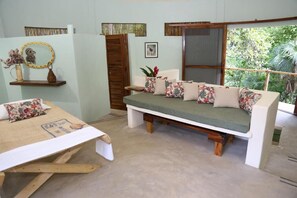 A room with a bed, a sofa, a window with a view of greenery, and a wall-mounted shelf with decorative items.