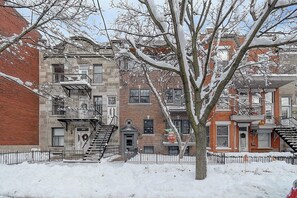 A row of brick townhouses with fire escapes, a tree in the foreground, and a snow-covered ground.