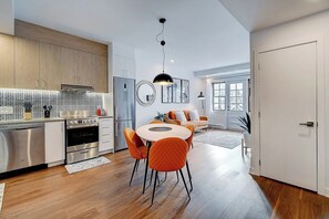 A modern kitchen with stainless steel appliances, a dining area with orange chairs, and a large window providing natural light.