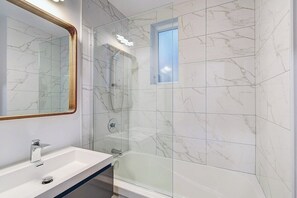 A bathroom with a white sink, a mirror with a wooden frame, a glass-enclosed shower, and marble tiles.