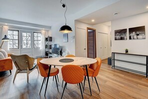 A modern dining area with a round table and orange chairs, a black pendant light, and a cityscape view.