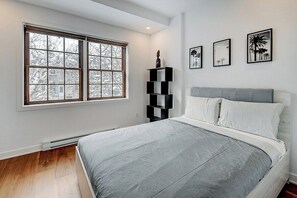 A bedroom with a large window, a bed with grey bedding, a black shelving unit, and three framed pictures on the wall.
