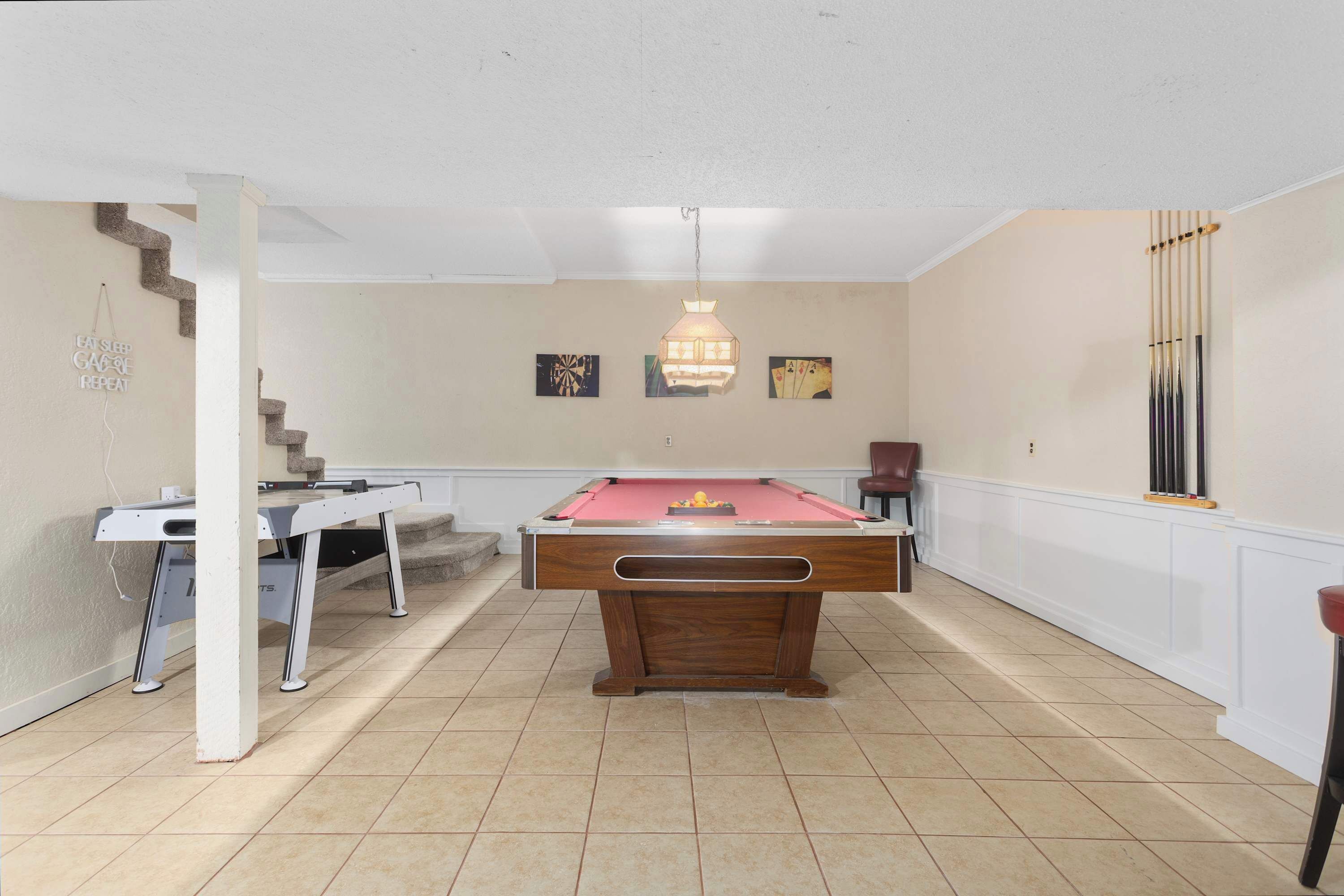 A pool table in a room with a staircase, a hanging light fixture, and a wall with framed artwork.