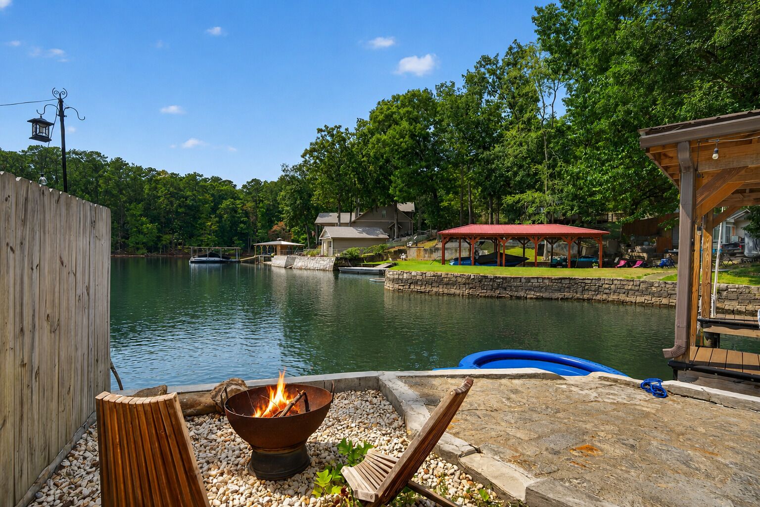 A lakeside outdoor area with a fire pit, a wooden deck, and a covered seating area.