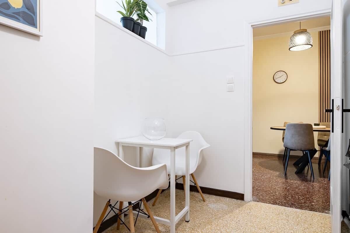 Um quarto pequeno com paredes brancas, uma mesa e duas cadeiras, um vaso de planta numa prateleira, e uma porta que dá para outro espaço.