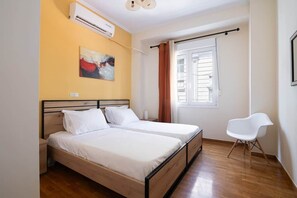 A bedroom with two beds, a window with curtains, a chair, and an air conditioning unit. - S homes | Athens · Retro-Modern 2BR · Attiki Metro (Athens)