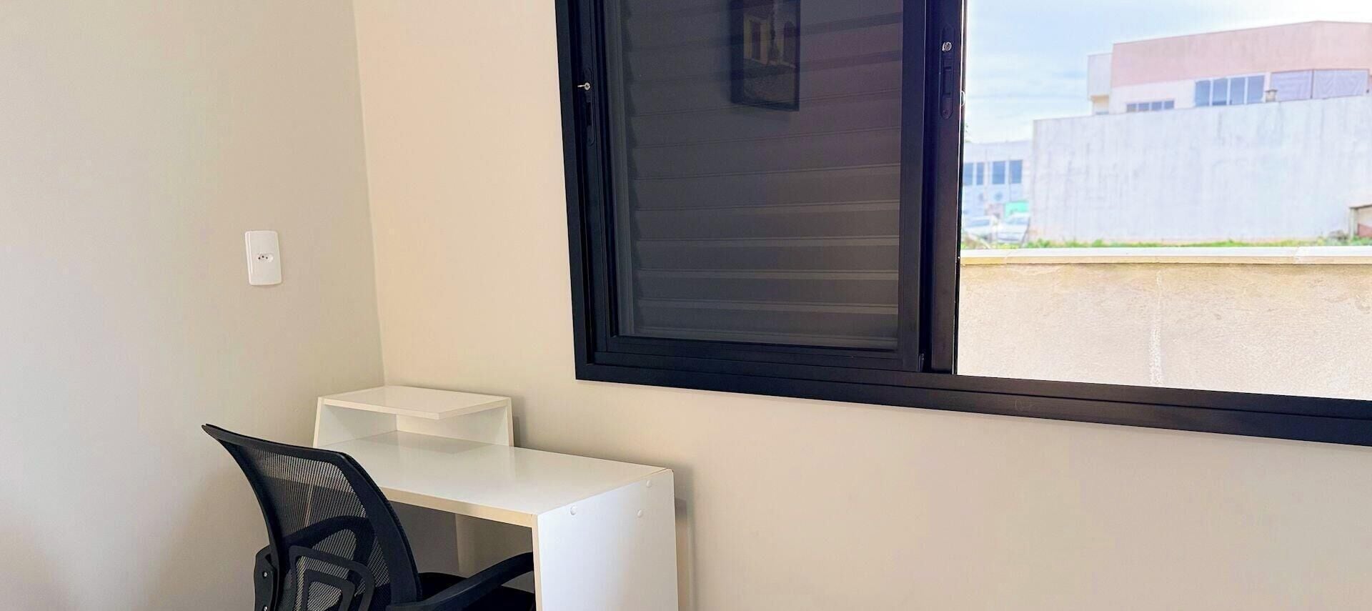 A room with a white desk, a black chair, a window with closed blinds, and a view of a building outside.