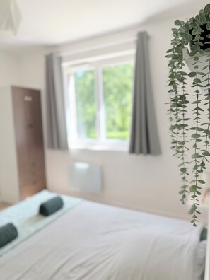 A bedroom with a bed, a window with curtains, a wall-mounted radiator, and a hanging plant. - Country house, garden with trees, 4 km from Fougères (Romagné)