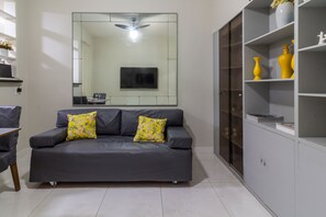 Ein modernes Wohnzimmer mit einem grauen Sofa, zwei gelben Kissen mit Blumenmuster, einem großen Spiegel, einem Deckenventilator und einer weißen Kommode mit gelben Vasen.