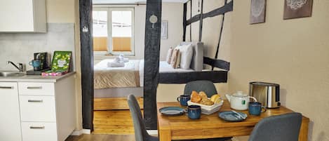 A cozy room with a wooden dining table, chairs, a bed with pillows, a window with blinds, and a kitchen area with a kettle and cups.