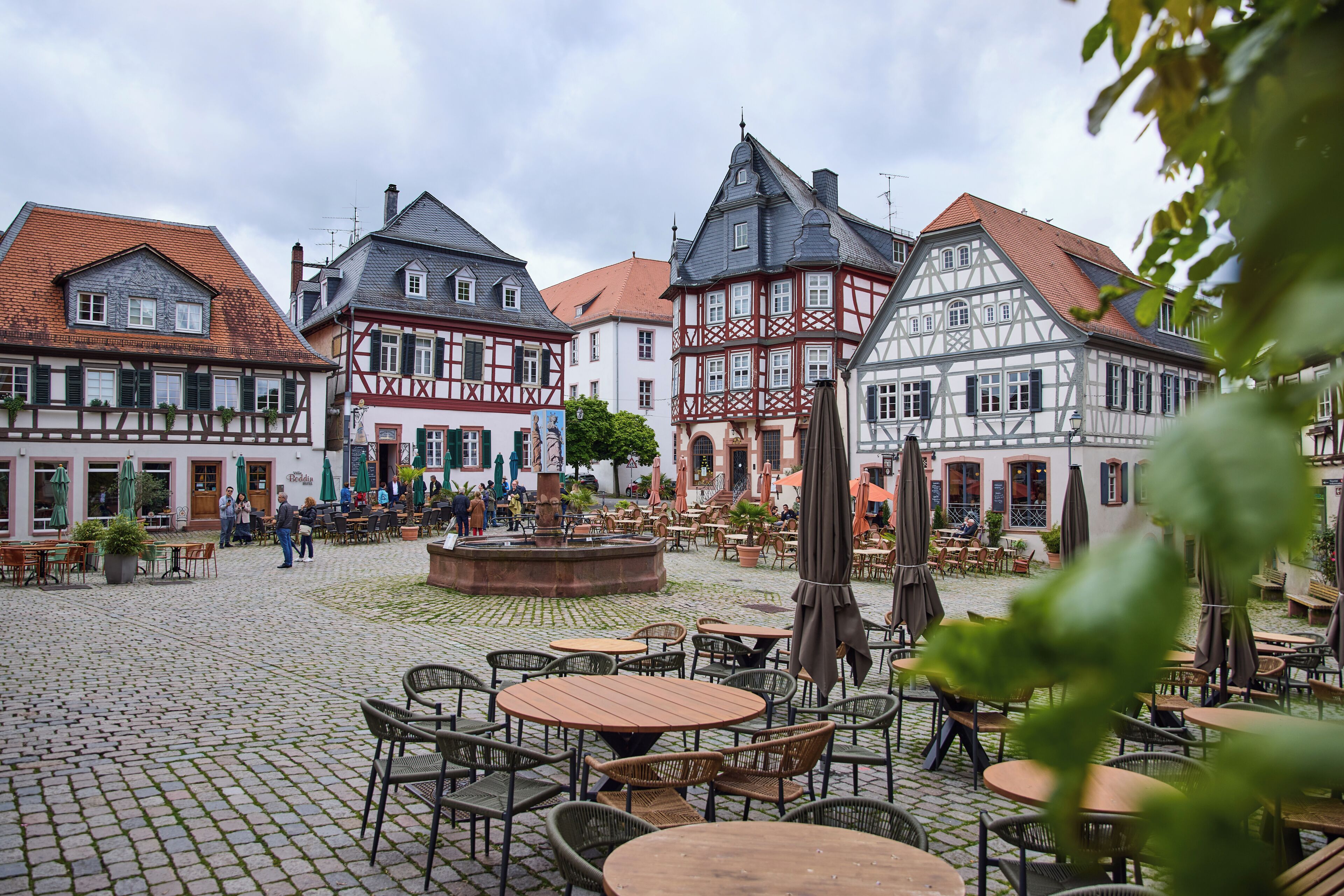 Ein Kopfsteinpflasterplatz mit traditionellen Fachwerkhäusern, Außenbestuhlung und einem zentralen Brunnen.