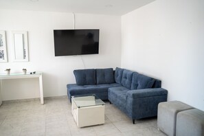 Ein Wohnzimmer mit einem Ecksofa, einem Glastisch, einem weißen Schreibtisch und einem wandmontierten Flachbildfernseher.