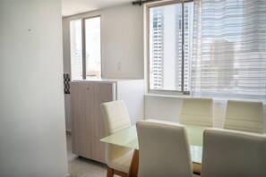 Ein Zimmer mit Esstisch und Stühlen, einem Fenster mit Vorhängen und Blick auf hohe Gebäude.