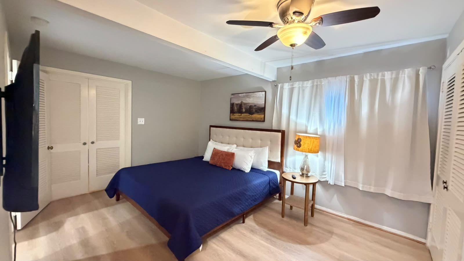Un dormitorio con una cama, un ventilador de techo, una mesita de noche con lámpara y una ventana con cortinas.
