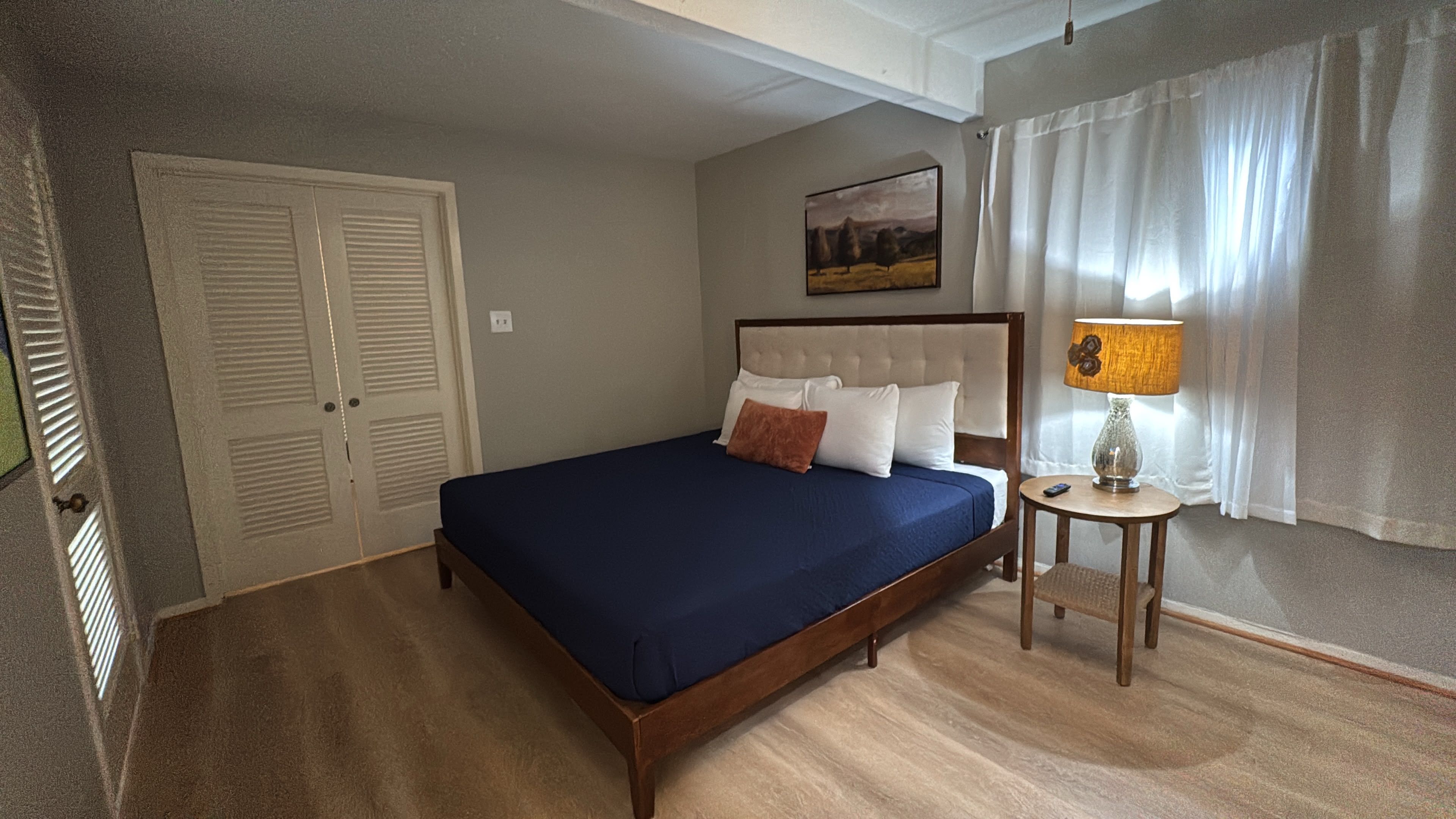 A bedroom with a bed, a bedside table with a lamp, a painting on the wall, and a door.