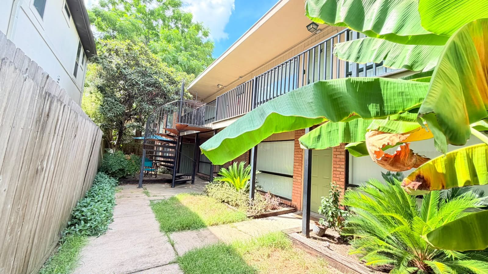 Quintal residencial com calçada pavimentada, cerca de madeira e diversas plantas, incluindo folhas de bananeira.