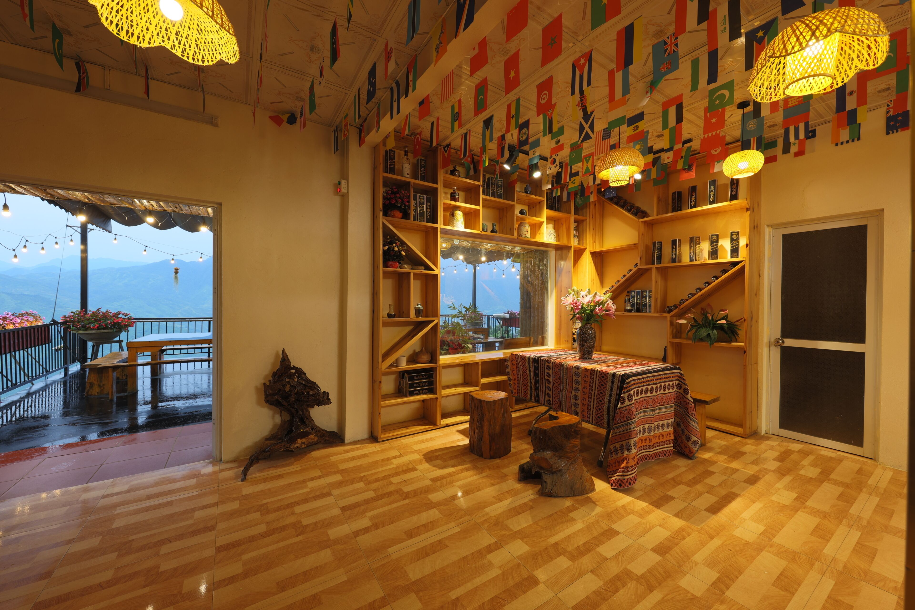 Une pièce avec un plancher en bois, une table recouverte d’une nappe à motifs, des étagères remplies de divers objets et une vue sur les montagnes par une grande fenêtre.