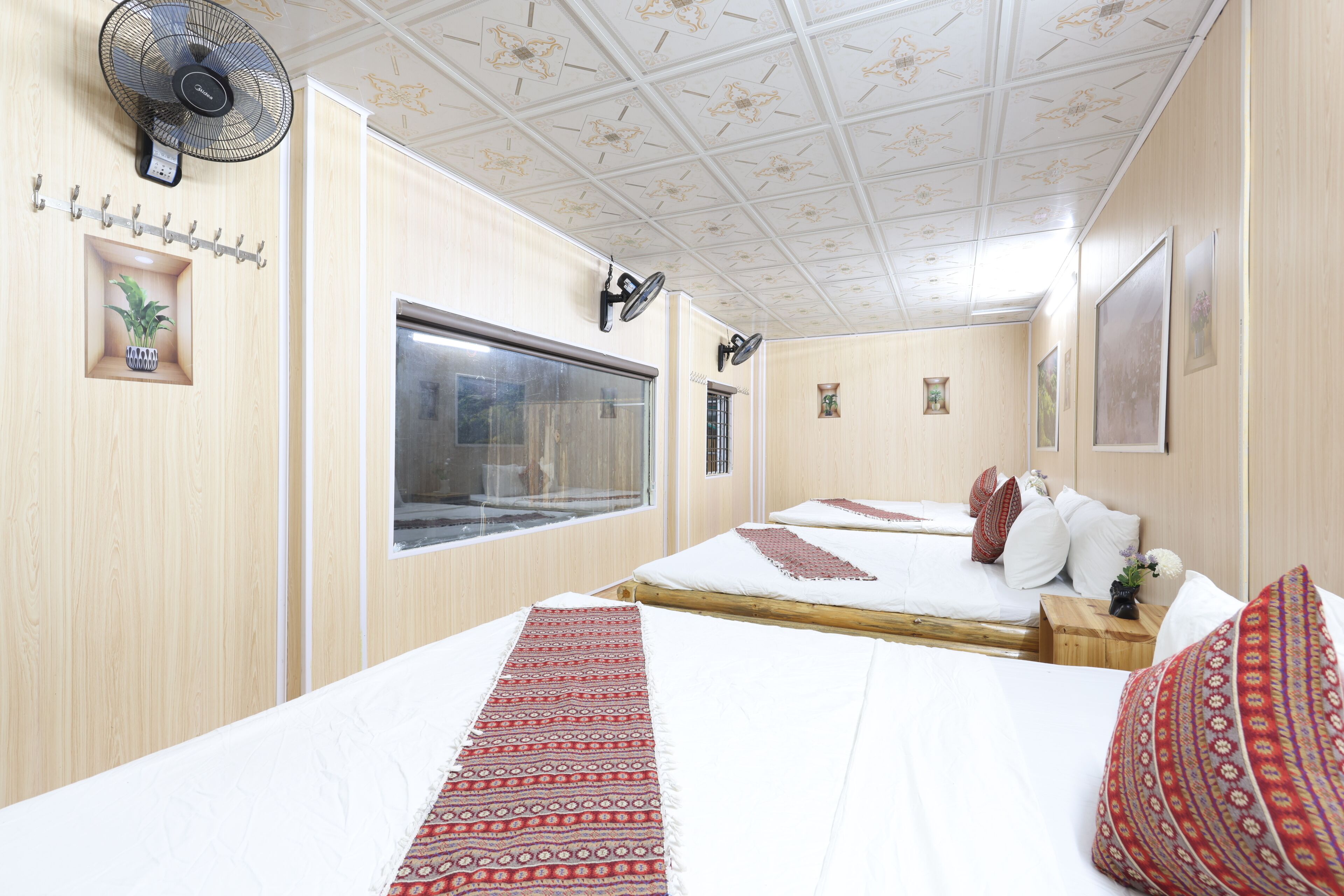 A hotel room with a large bed, a smaller bed, a window, a ceiling fan, and a wall-mounted light.