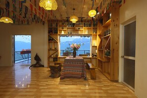 Ein Zimmer mit Holzböden, einem zentralen Tisch mit gemustertem Tischläufer, Regalen mit Büchern und Dekorationsgegenständen sowie Blick auf Berge durch ein Fenster.