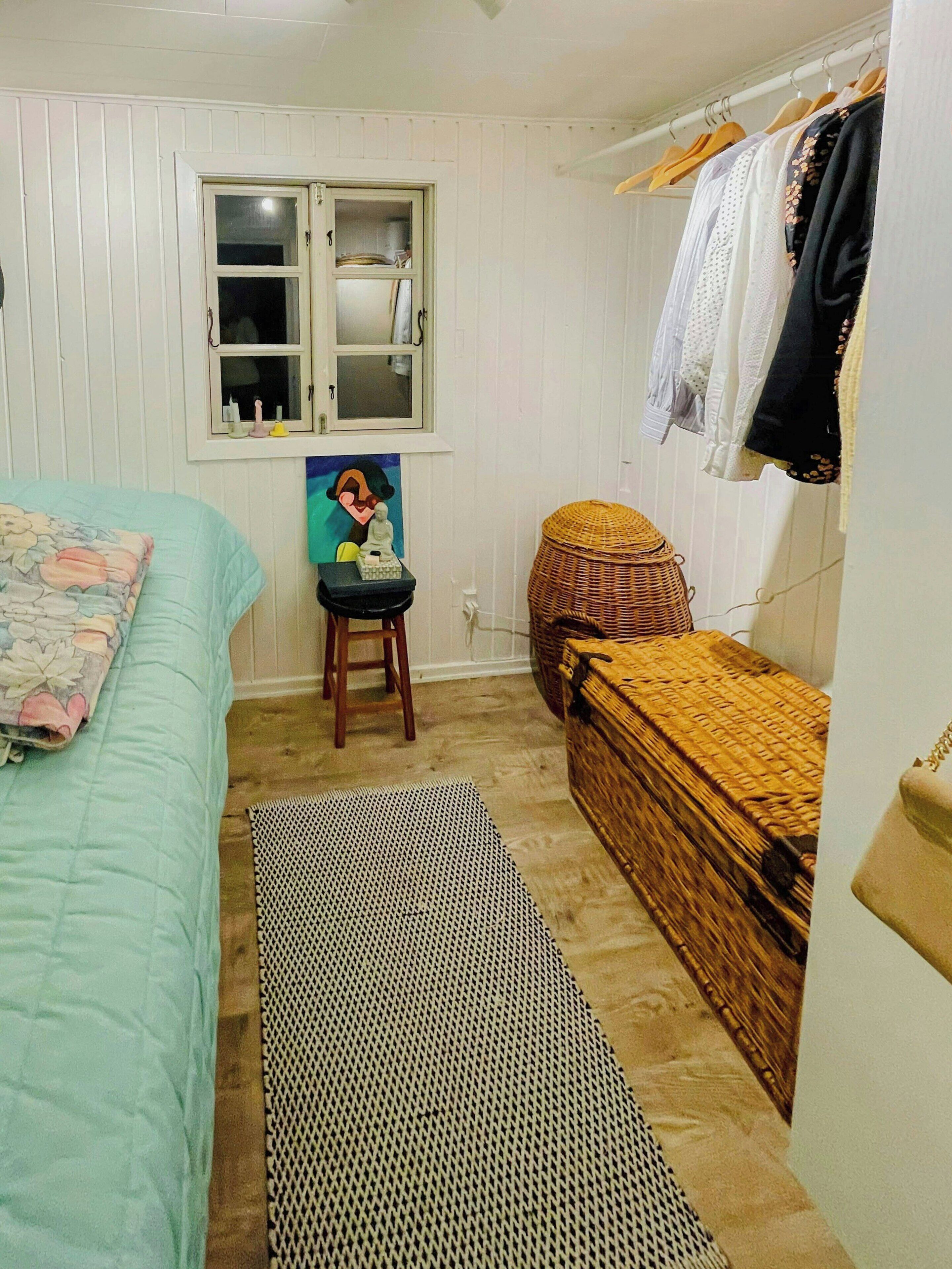 Ein Schlafzimmer mit einem Bett, einem kleinen Hocker, einer Korbschrank, einer Bank, einem Fenster und einem Kleiderständer.
