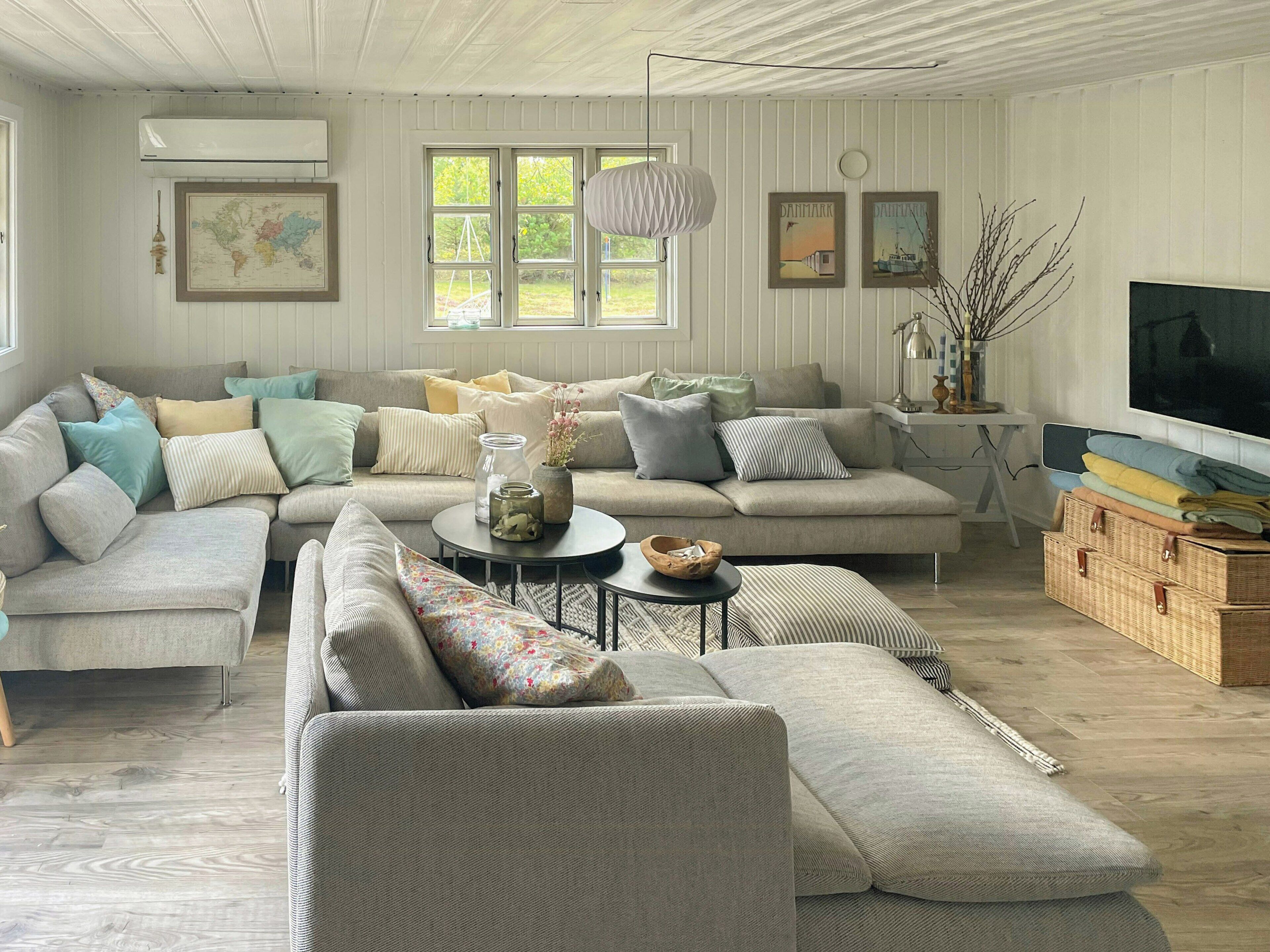 En stue med en hjørnesofa, et sofabord og et fjernsyn. Der er vinduer med gardiner, et kort på væggen og en lampe.