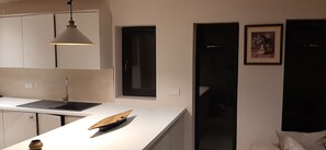 Eine moderne Küche mit weißer Arbeitsplatte, einer hängenden Pendelleuchte, einem Fenster mit schwarzem Rahmen und einem Bilderrahmen an der Wand.