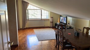 Ein Zimmer mit Esstisch, Stühlen, Fernseher, Heizkörper und einem fensterbelaubten Fenster.