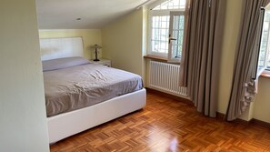 Ein Schlafzimmer mit Bett, Nachttisch, Lampe, einem Fenster mit Vorhängen und einer Heizung.