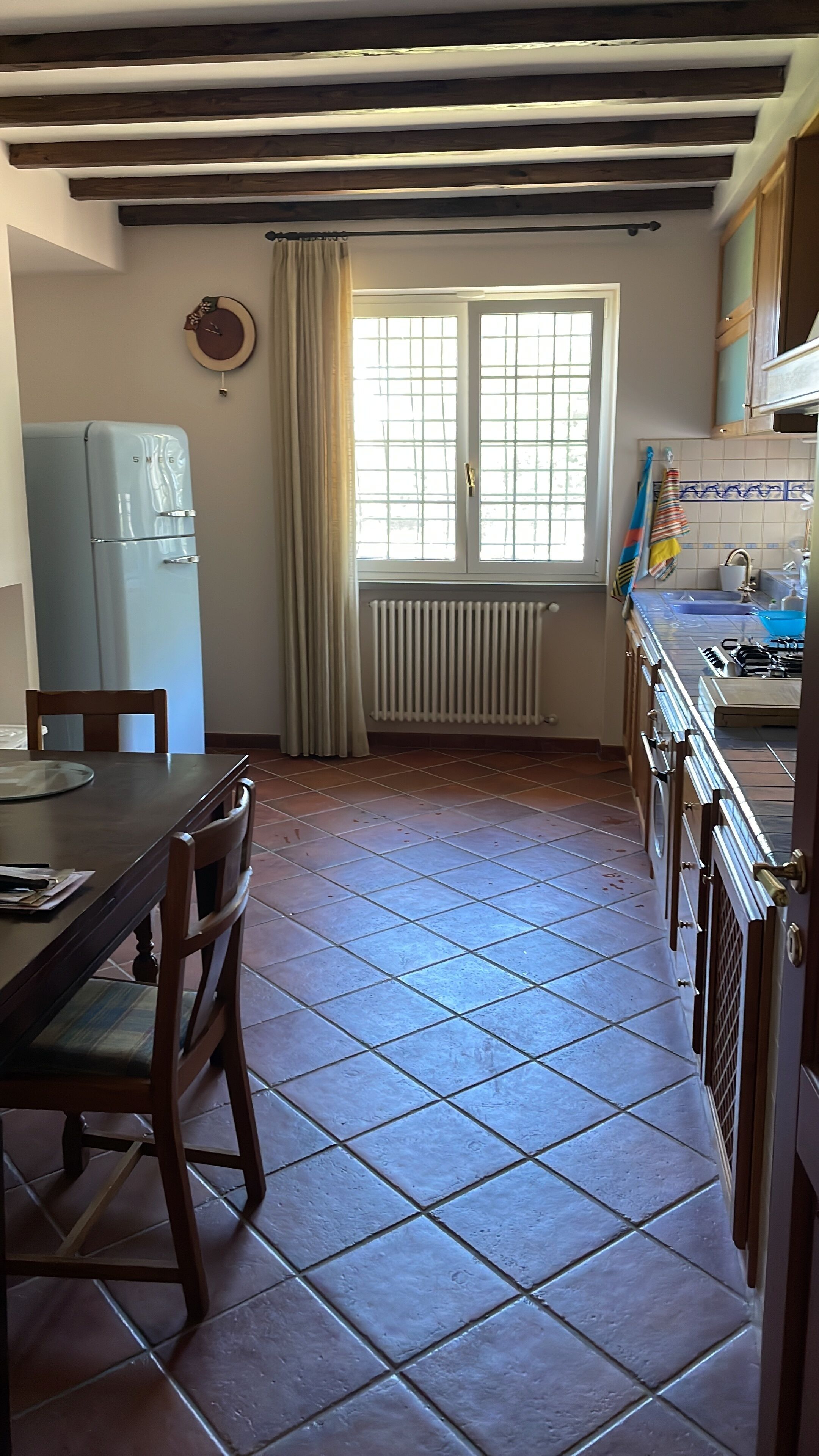Een keuken met een eettafel, stoelen, een koelkast, een raam met gordijnen en een betegelde vloer.