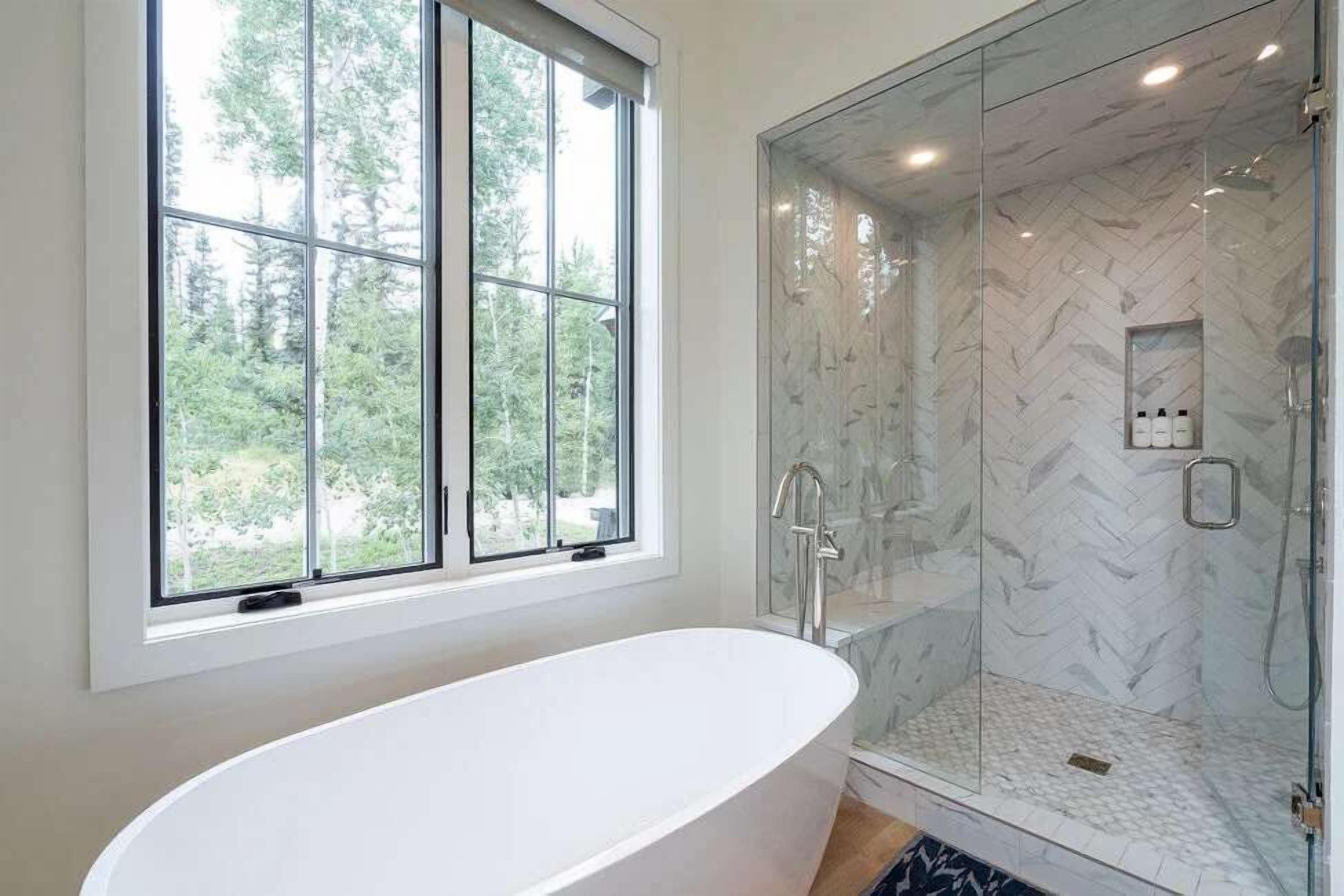 A modern bathroom with a freestanding bathtub, a glass-enclosed shower, and a window providing a view of trees.
