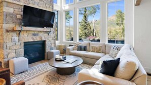 A living room with a stone fireplace, a flat-screen TV mounted above it, a beige sofa, and a coffee table. - Luxe Telluride Oasis - Hot Tub & Deck & Views (Mountain Village)