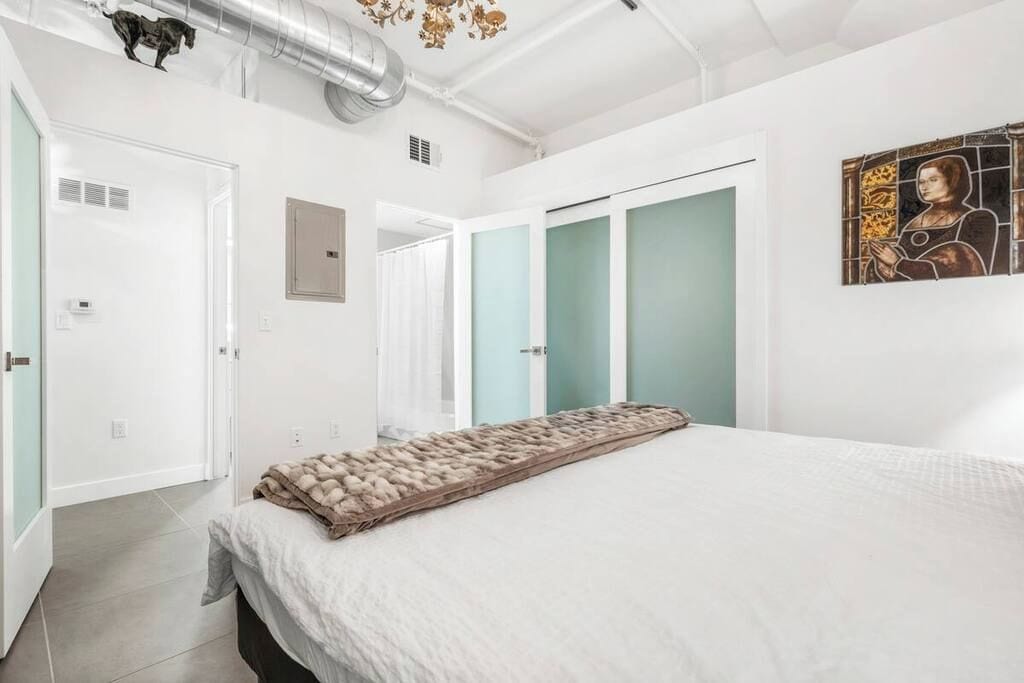 A bedroom with a bed, a door, a wall-mounted artwork, and a ceiling vent.