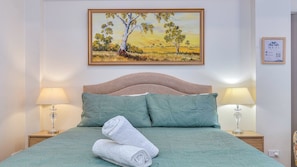 A neatly made bed with a green bedspread and two pillows, a painting of a landscape above it, and two bedside lamps on either side. - Berkeley Studio by HolidayCo. (BERKELEY VALE)