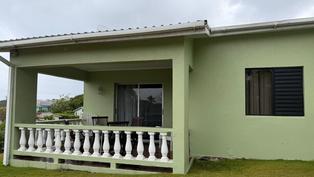Ein eingeschossiges Gebäude mit grüner Fassade, einer Veranda mit weißen Balustraden und einem Fenster mit schwarzen Rollläden.