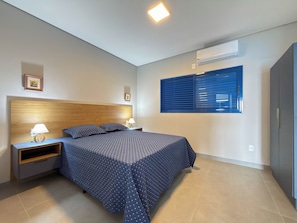 A bedroom with a bed, nightstands, a wall-mounted air conditioner, and a window with blinds.