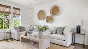 A modern living room with a white sofa, a coffee table, and a side table with a lamp. There are woven wall decorations and a view of greenery outside. - Sully & Sonshine | 3 BR Near Sullivan's Island (Mount Pleasant)