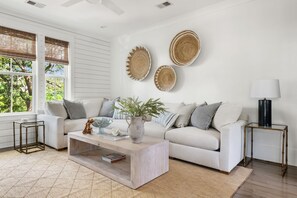 A modern living room with a white sofa, a coffee table, and a side table with a lamp. There are woven wall decorations and a view of greenery outside. - Sully & Sonshine | 3 BR Near Sullivan's Island (Mount Pleasant)
