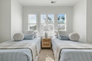 A bedroom with two beds, a nightstand, and a window with blinds. - Sully & Sonshine | 3 BR Near Sullivan's Island (Mount Pleasant)