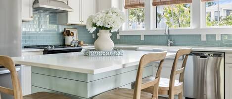 Una cocina moderna con armarios blancos, una isla central con taburetes y luces colgantes, además de un lavavajillas.
